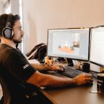Homem usando fones de ouvido trabalha em um computador com dois monitores em um escritório moderno.
