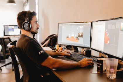 Homem usando fones de ouvido trabalha em um computador com dois monitores em um escritório moderno.