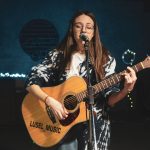 Cantora tocando violão e cantando ao microfone em um palco.