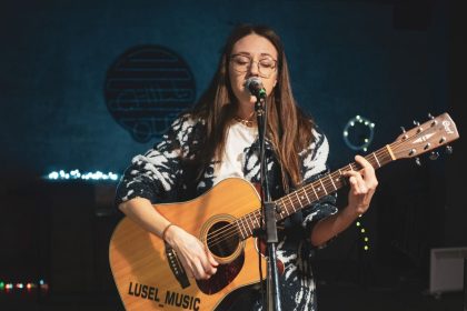 Cantora tocando violão e cantando ao microfone em um palco.