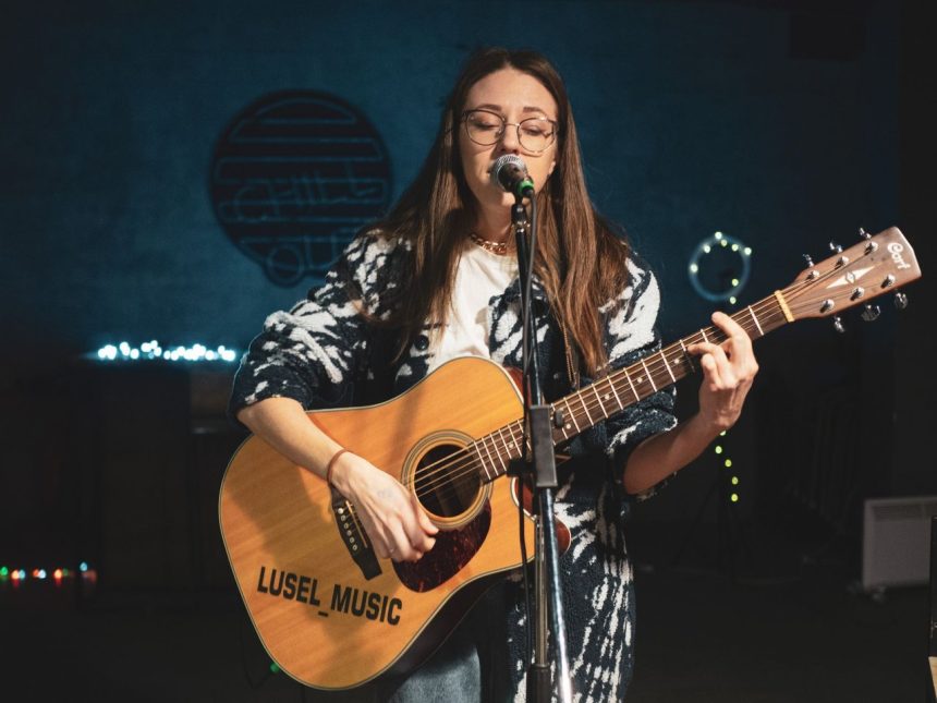 Cantora tocando violão e cantando ao microfone em um palco.