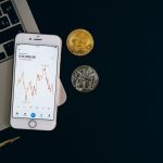 Smartphone exibindo um gráfico de preços de Bitcoin ao lado de moedas de Bitcoin e Ethereum sobre fundo preto.