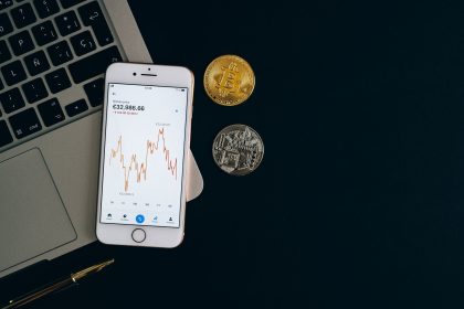 Smartphone exibindo um gráfico de preços de Bitcoin ao lado de moedas de Bitcoin e Ethereum sobre fundo preto.