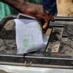 Mão colocando uma cédula de papel em uma urna eleitoral transparente ao ar livre